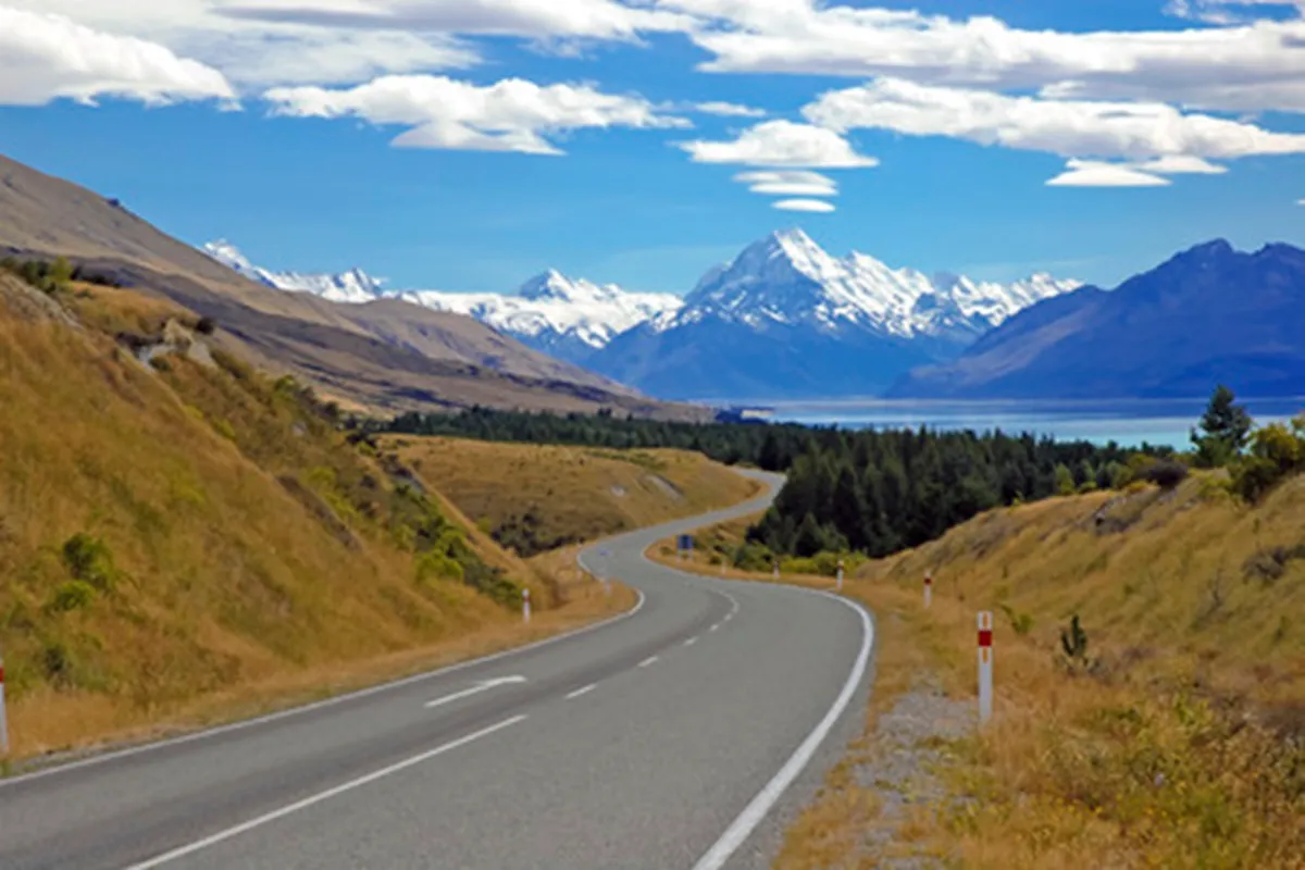 Winding sealed road through golden tussock hills towards a snow-capped mountain and a blue lake under a bright sky.