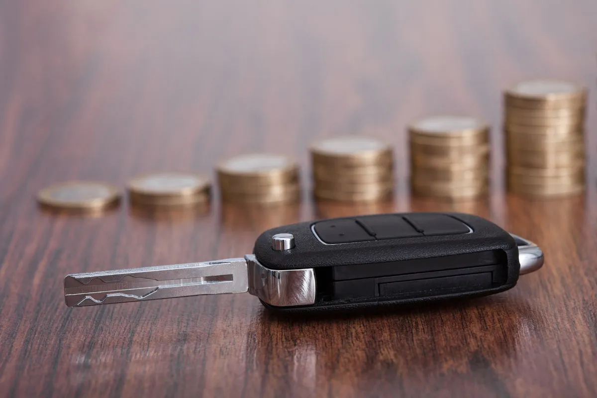 Car key lying on a wooden table in front of rising stacks of coins.
