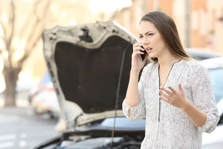 Woman on a phone call looking stressed beside a car with its bonnet raised.