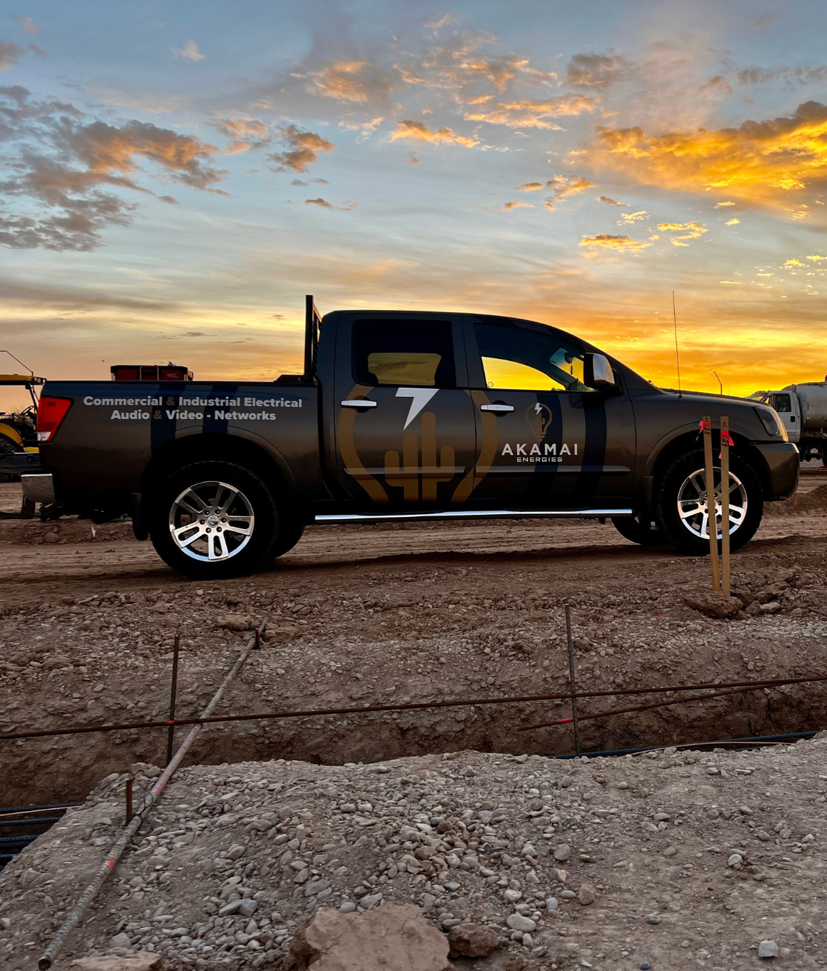 Black Akamai Energies pickup truck with company logo and text about commercial and industrial electrical services parked on dirt at sunset.