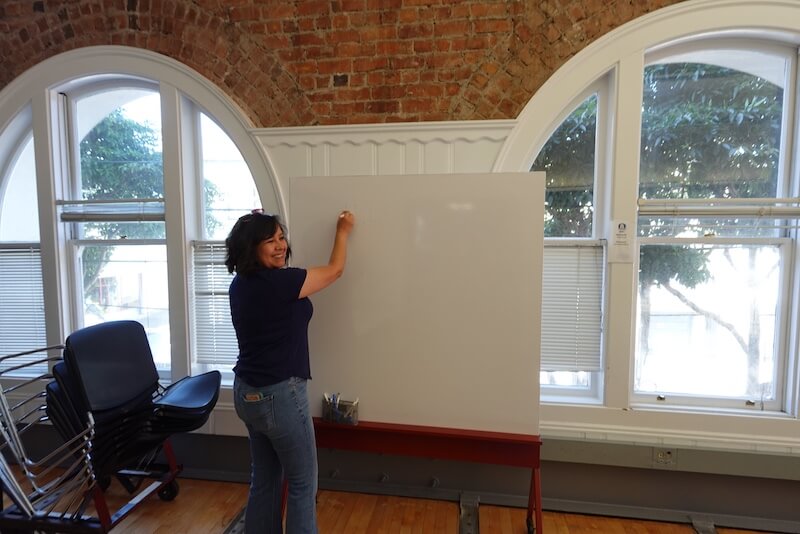 Cinthia Carvajal smiling and standing at a whiteboard inside a sunlit room with arched windows and exposed brick at The Women’s Building.