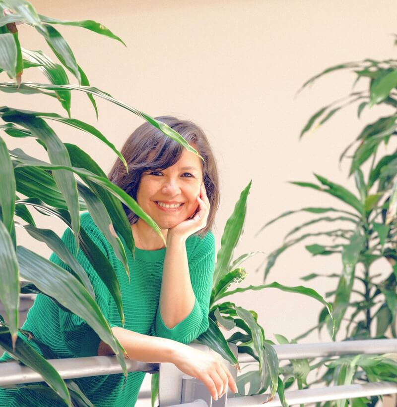 Cinthia Carvajal smiles warmly while leaning on a railing, surrounded by green plants in a sunlit space.