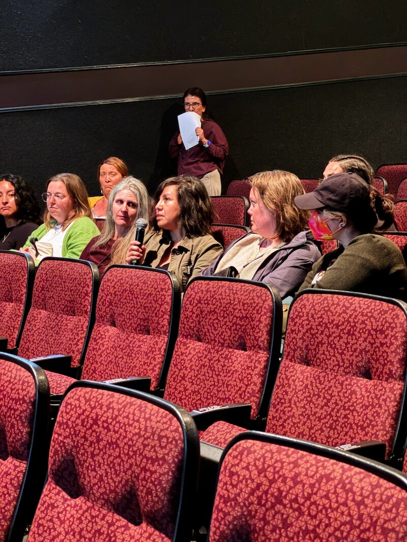 Cinthia Carvajal holds a microphone while speaking among a group of women seated in a theater, during a community conversation.