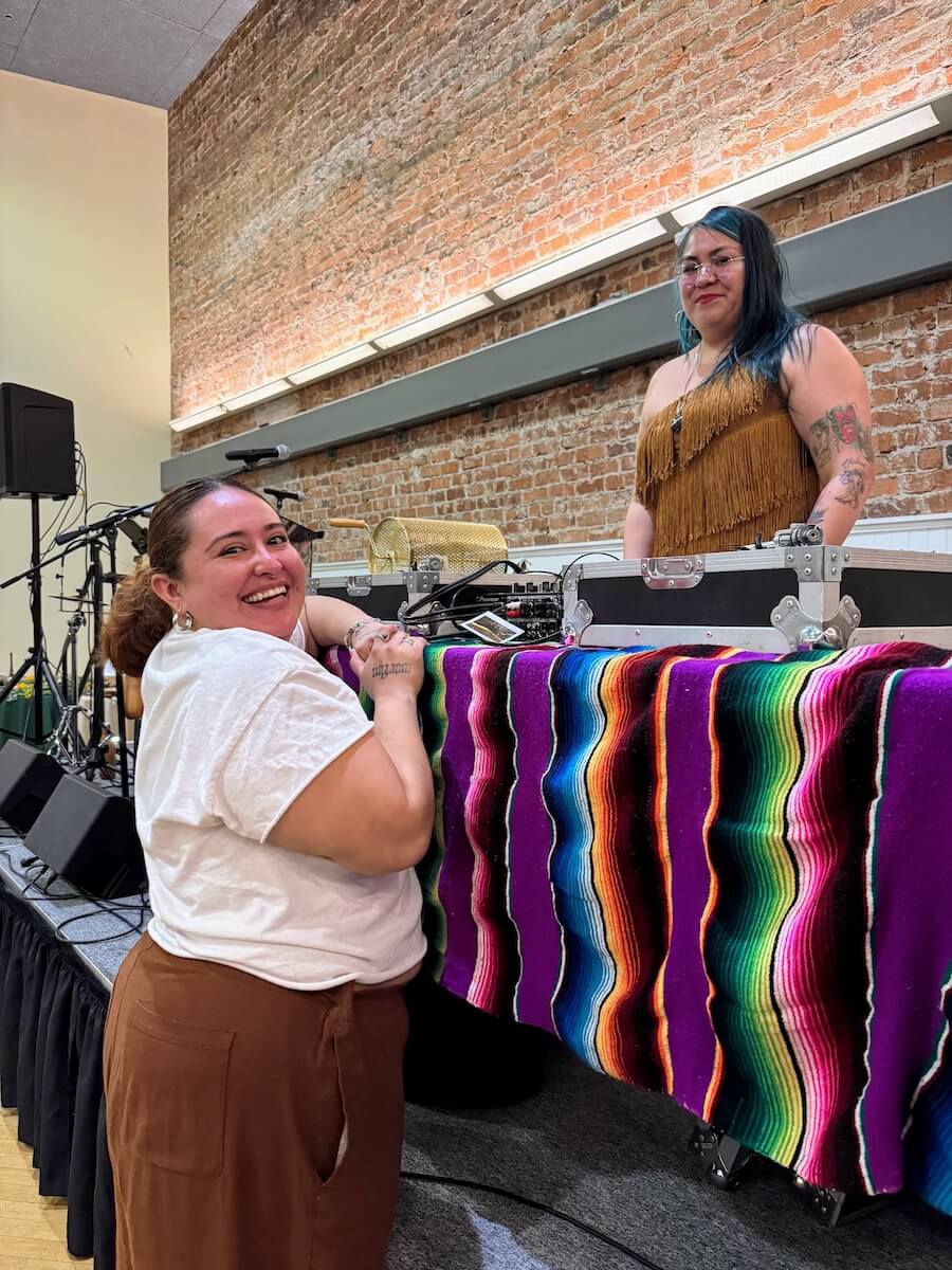 Vanessa Haro smiles beside a DJ table during a community event at The Women’s Building, with colorful textiles and sound equipment in the background.