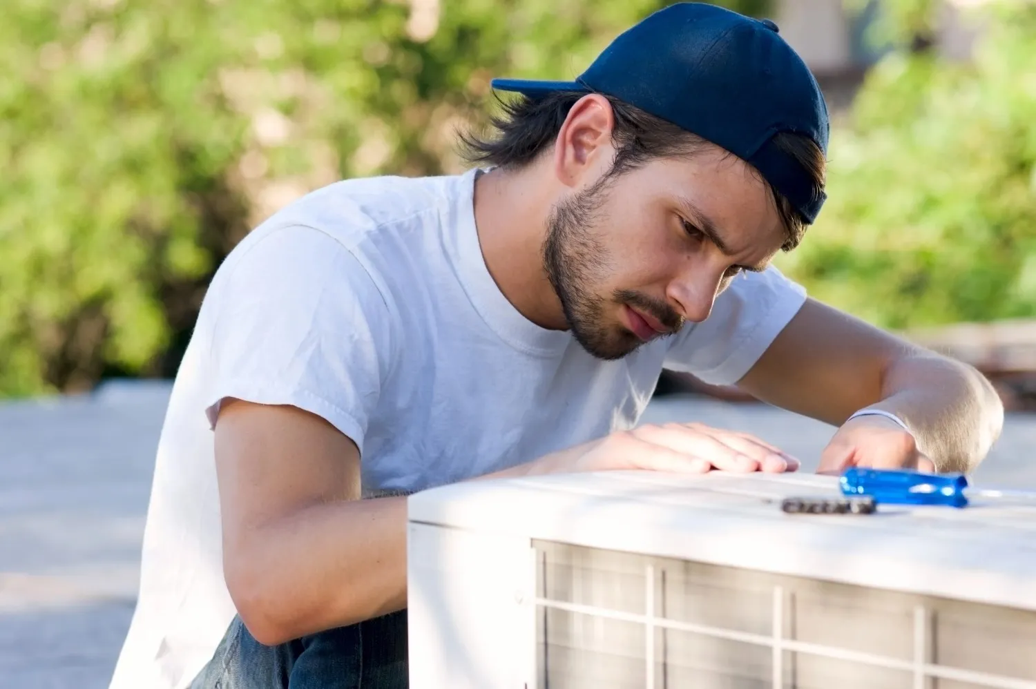 A technician wearing a tool belt and a cap is kneeling and repairing an outdoor heat pump unit next to a modern wooden wall