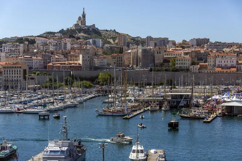 Vue du Vieux-Port de Marseille avec des bateaux amarrés et la basilique Notre-Dame de la Garde sur une colline en arrière-plan par une journée ensoleillée.