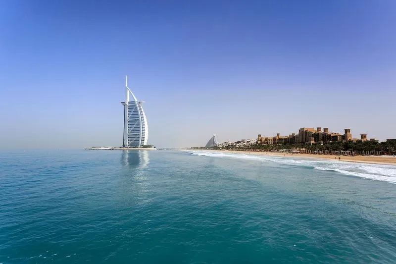 Vue sur la tour Burj Al Arab blanche en forme de voile face à la mer bleue à Dubaï avec une plage et des bâtiments au loin sous un ciel bleu clair.