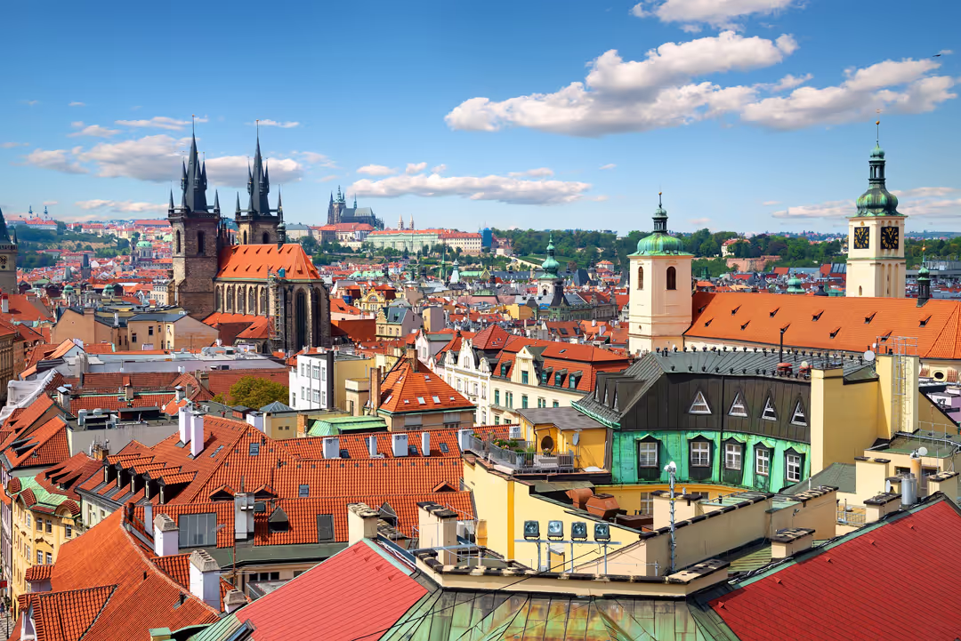 Vue aérienne de Prague avec ses toits en tuiles rouges, églises gothiques et le château de Prague en arrière-plan sous un ciel bleu avec quelques nuages.