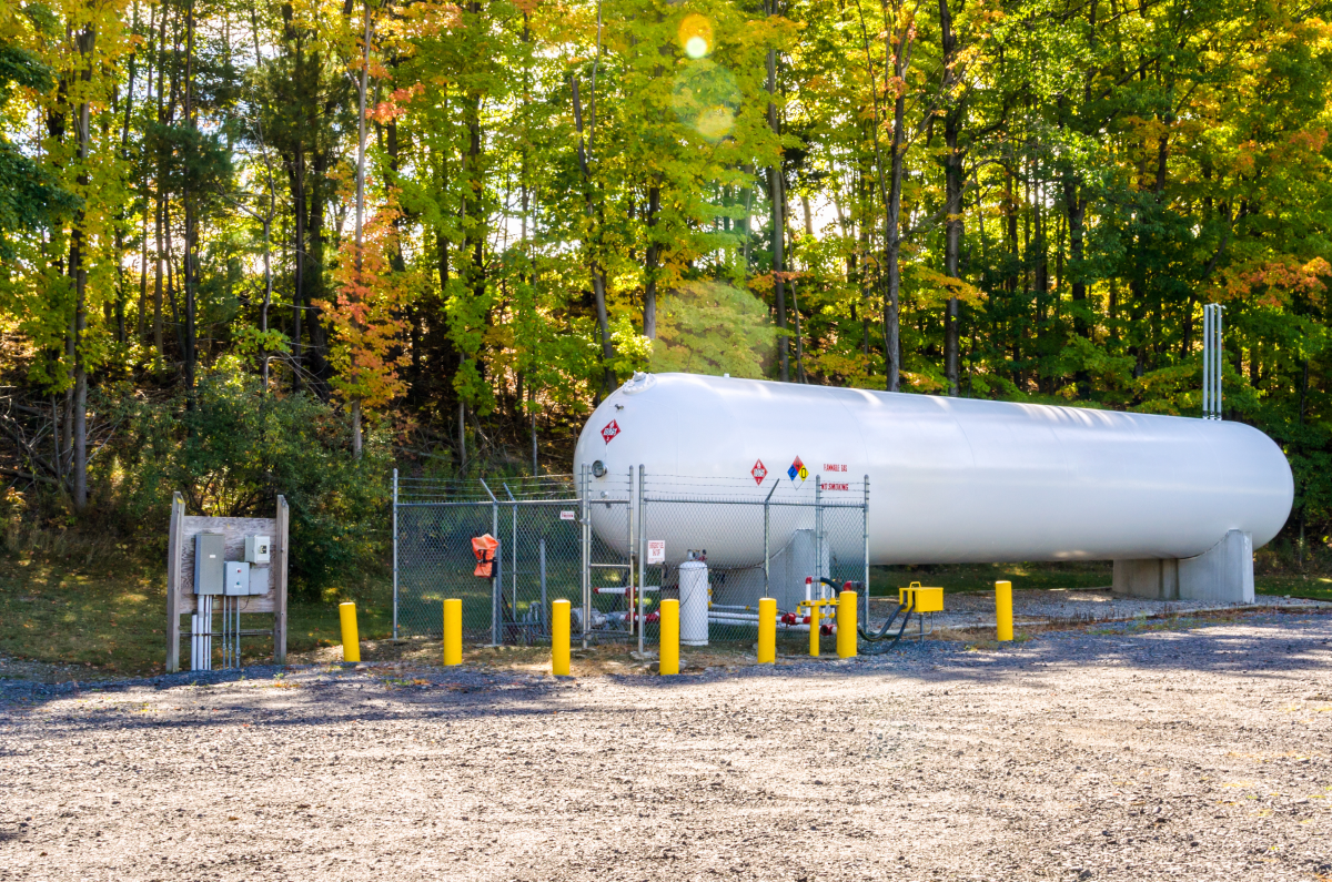 Large white flammable gas storage tank enclosed by a chain-link fence with yellow protective posts, set against a background of green and yellow trees.