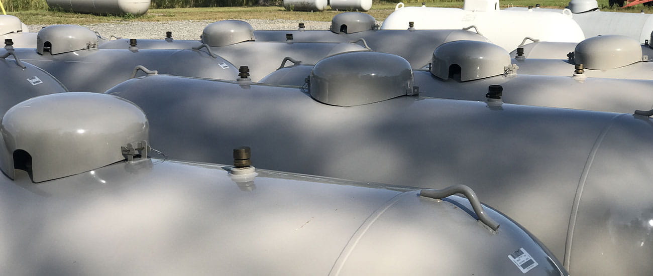 Cluster of large gray propane tanks outdoors on gravel with grass in the background.