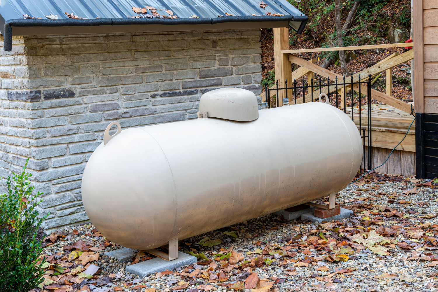 Large beige propane tank installed on a gravel bed beside a stone and wood house with fallen autumn leaves around.