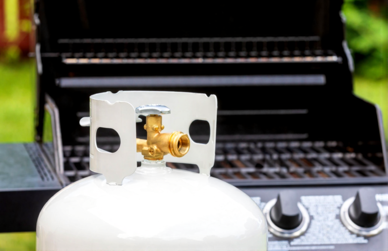 Close-up of a white propane tank valve in front of a black outdoor gas grill with control knobs.