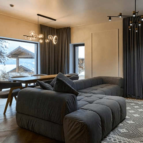 Modern living room with a large dark grey sectional sofa, wooden dining table, large windows with dark curtains, and contemporary lighting fixtures.