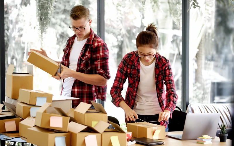 En man och en kvinna iklädda rödsvart rutiga skjortor som står vid ett bord fyllt av små kartonger som håller på att packa ordrar. 