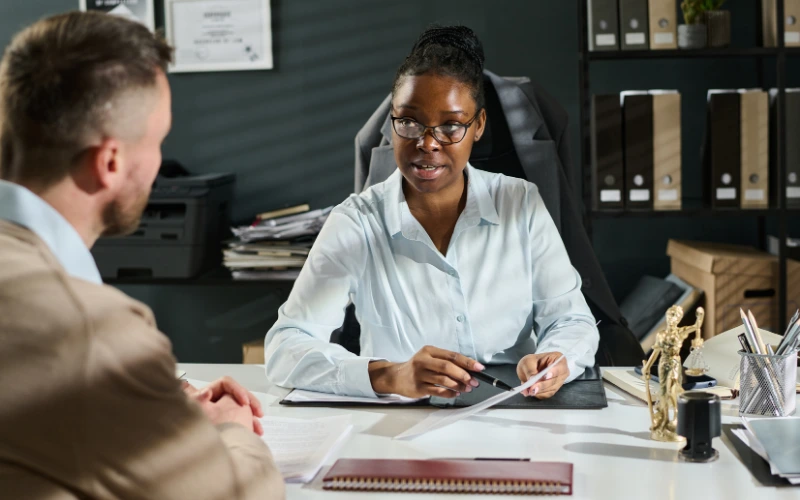 En kvinna i blå skjorta sitter i ett kontor vid ett skrivmord med en man mittemot i kostym där hon förklarar något för honom.