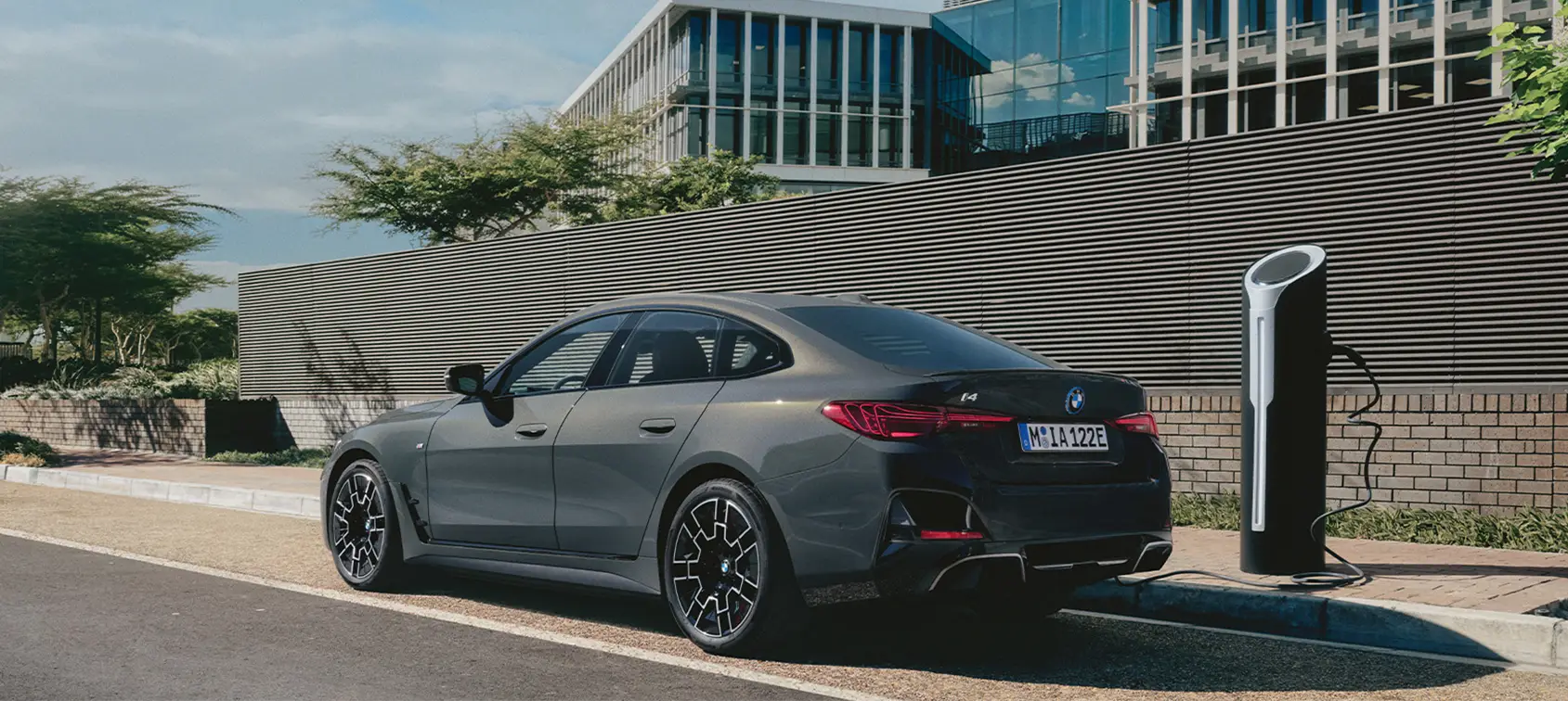 BMW i4 in mattem Grau beim Laden an moderner E-Ladesäule vor urbaner Glasarchitektur – BMW Köhler in Karlstadt