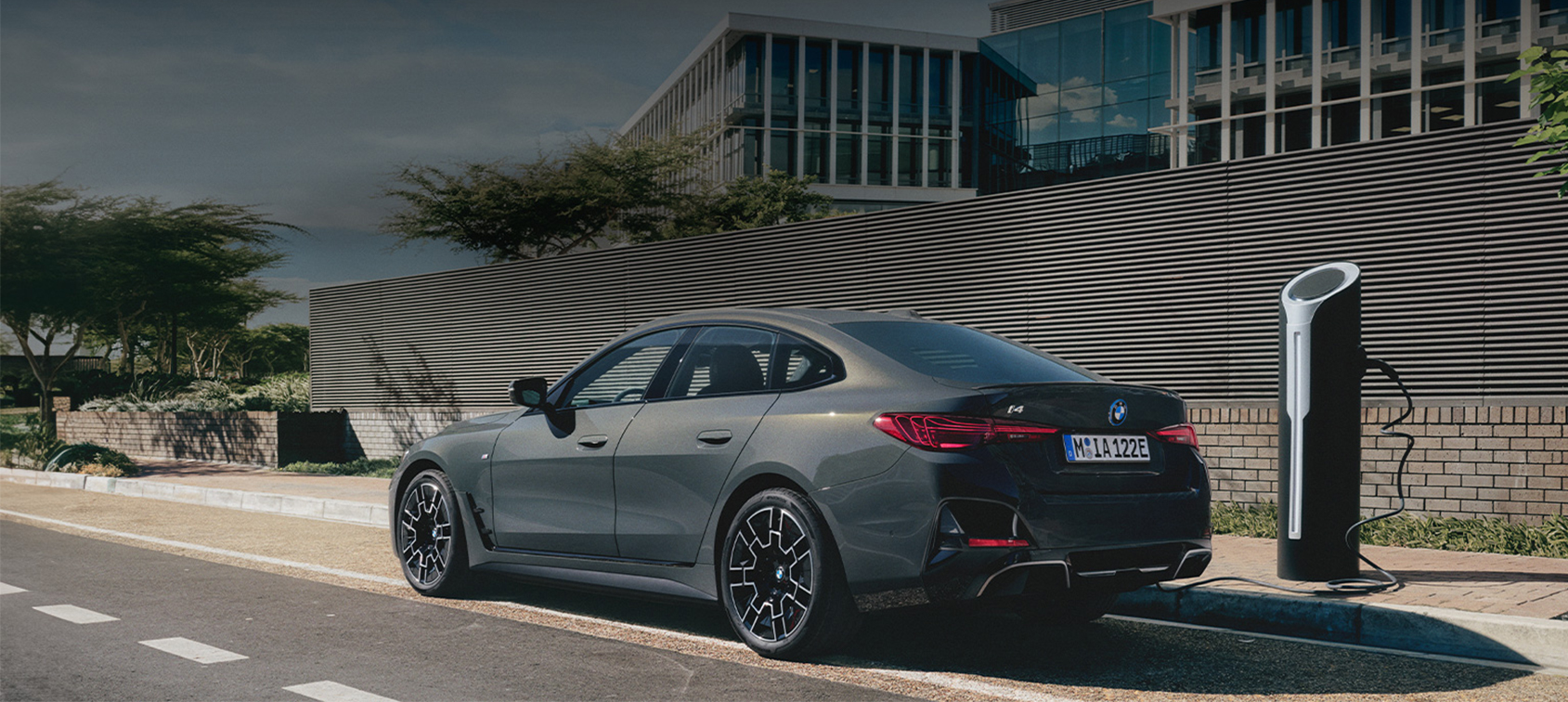 BMW i4 in mattem Grau beim Laden an moderner Ladesäule vor urbanem Bürogebäude – BMW Köhler in Karlstadt