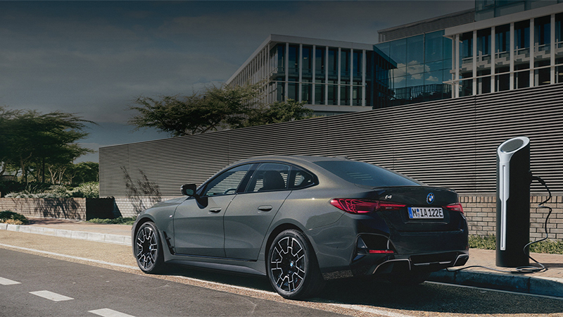 BMW i4 in mattem Grau beim Laden an moderner Ladesäule vor urbanem Bürogebäude – BMW Köhler in Karlstadt