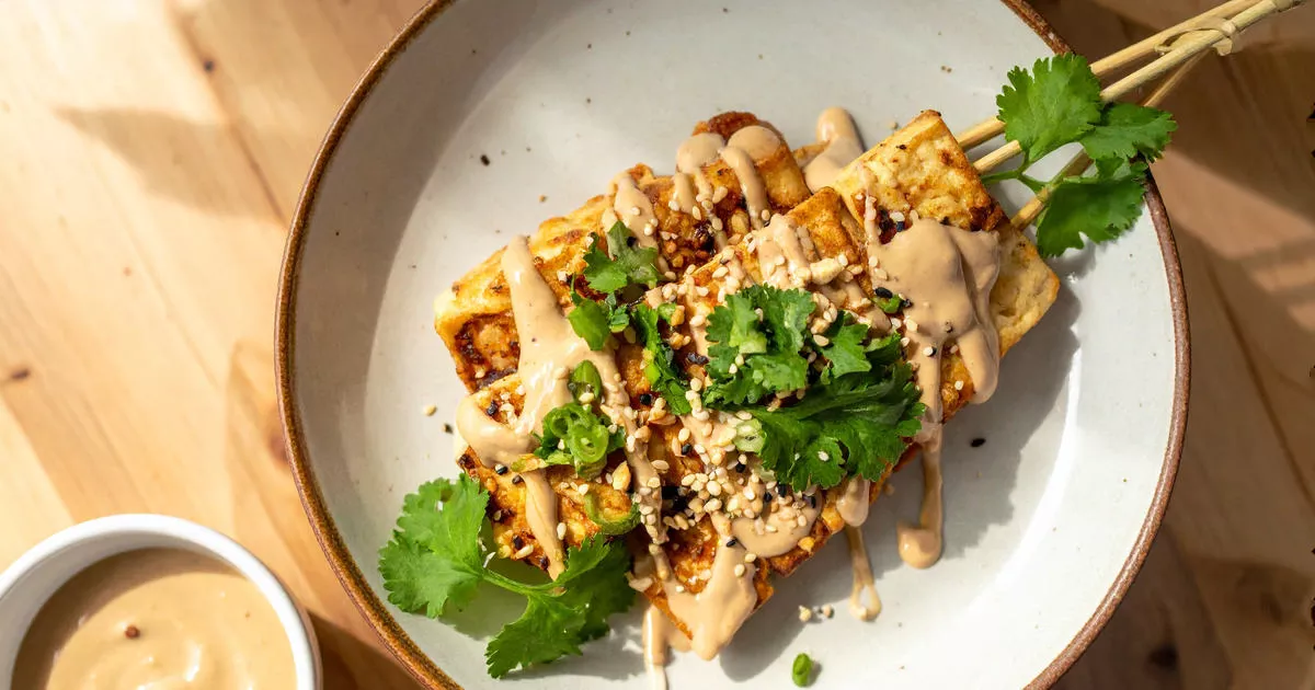 Tofu-Spieße mit Erdnusssauce, garniert mit Koriander, Sesam und Frühlingszwiebeln, serviert auf einem Holzbrett