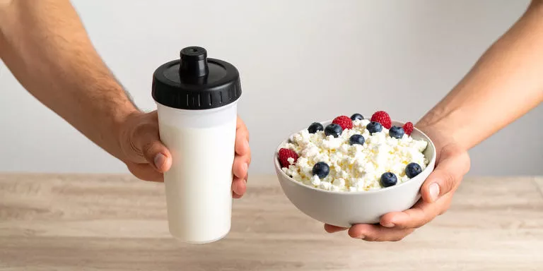 Zwei Hände halten je einen Snack – einmal ein Proteinshake, einmal eine natürliche Eiweißquelle mit Hüttenkäse und Beere