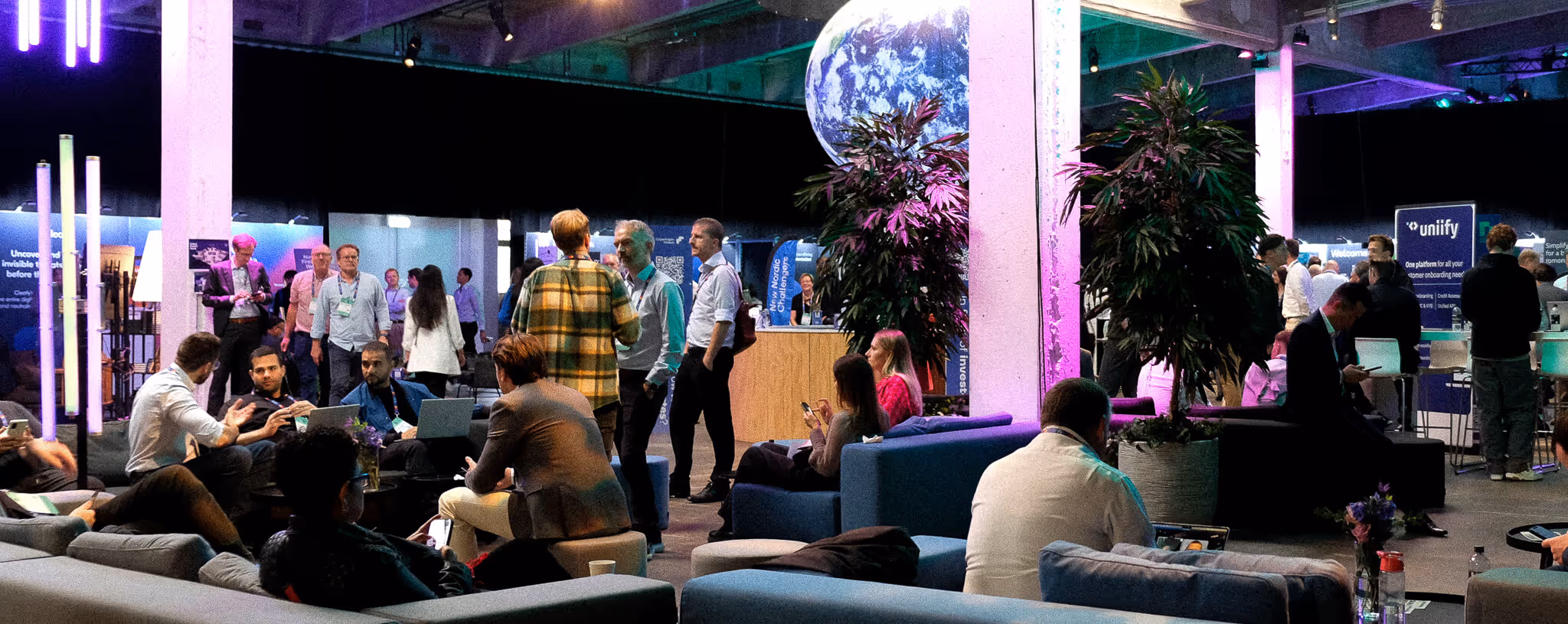 People networking and working on laptops in a modern conference lounge with ambient purple lighting and a large Earth globe decoration. On-site name badges