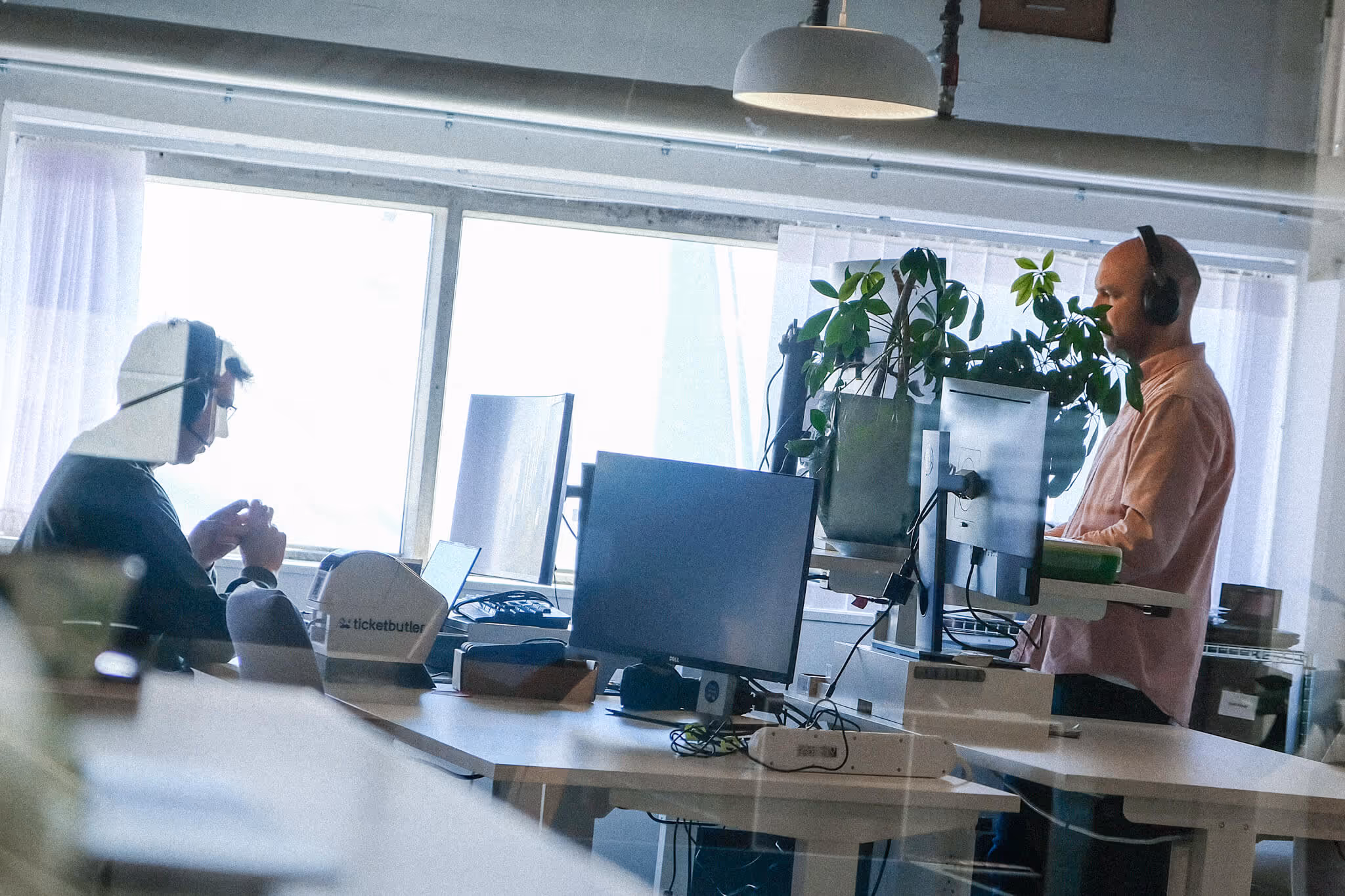 Two men wearing headsets working at separate computer desks in a bright office with large windows and plants.