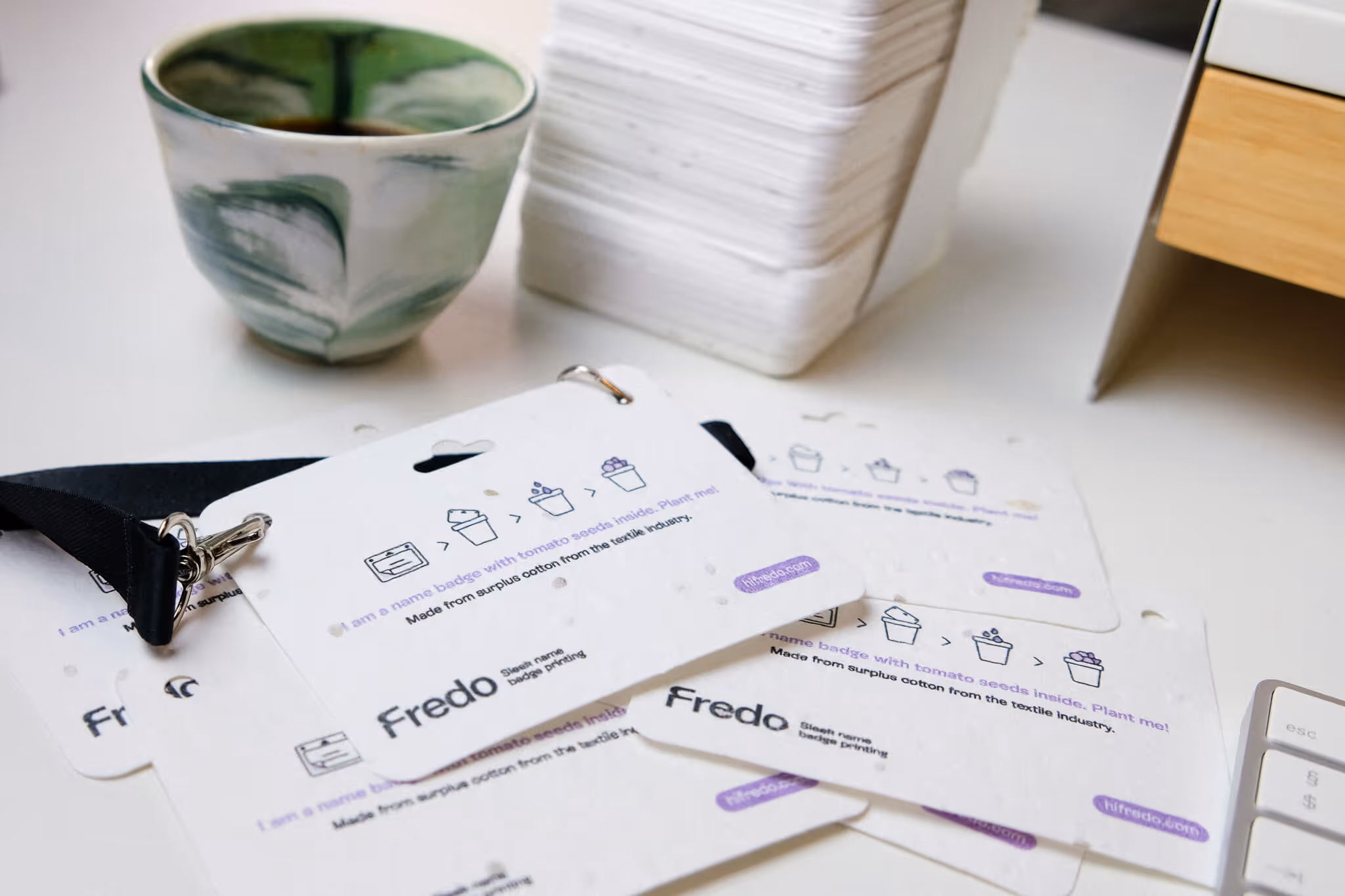 Plantable name badges with tomato seeds inside, printed with the name Fredo, arranged on a white desk near a green ceramic cup and a stack of white cards.