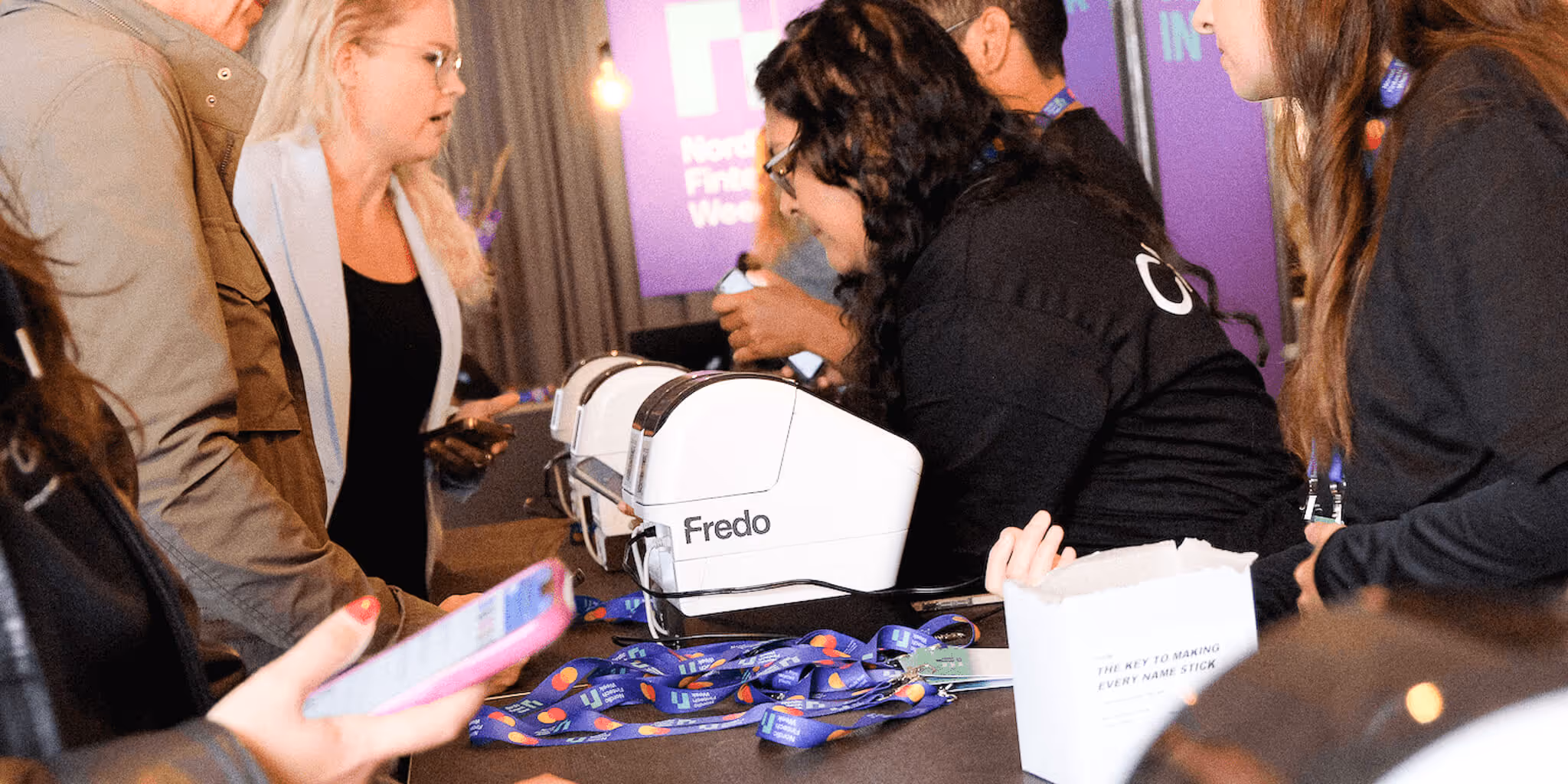 People interacting around a table with name badge printers labeled Fredo and blue event lanyards scattered on it.