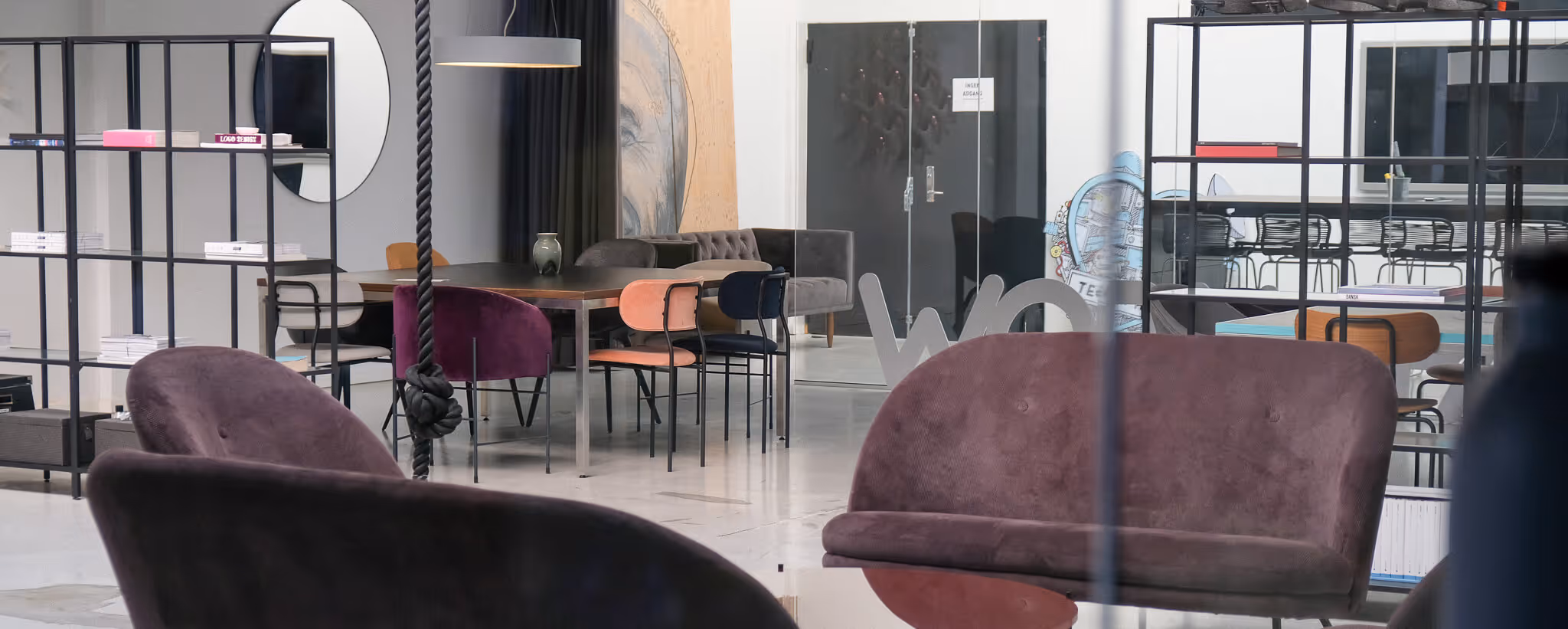 Modern office lounge area with purple velvet chairs, a table surrounded by colorful chairs, and shelving units holding books and decor.