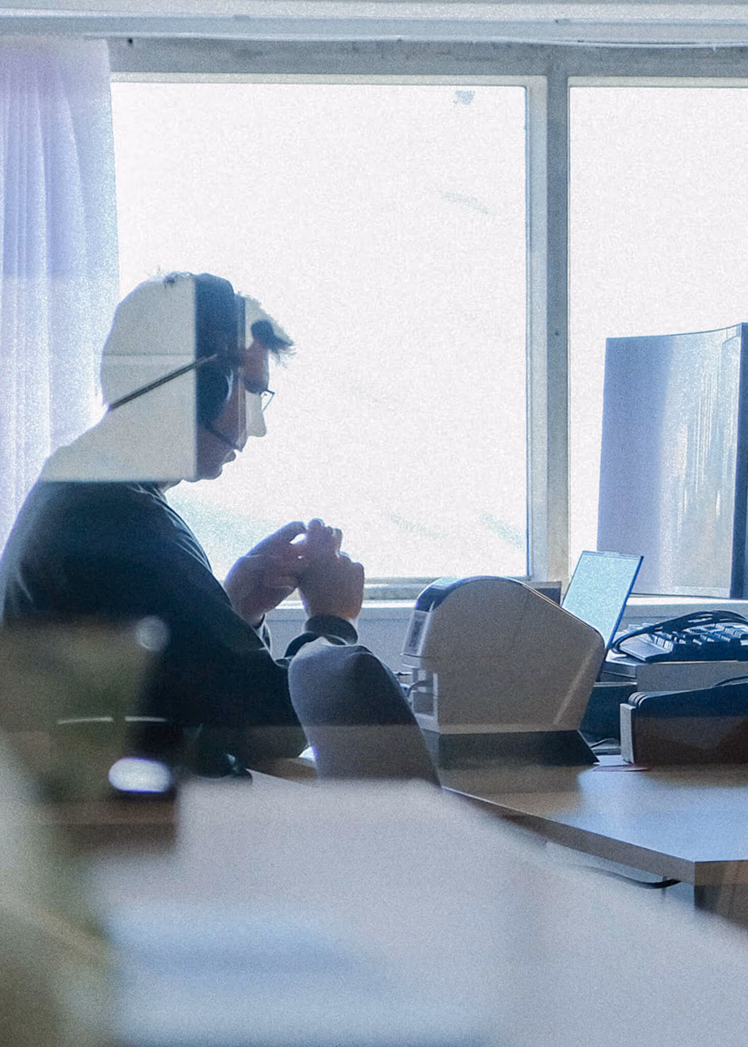 Silhouette of a person wearing glasses and a headset, sitting at a desk with office equipment near a large window. support phone. Global support