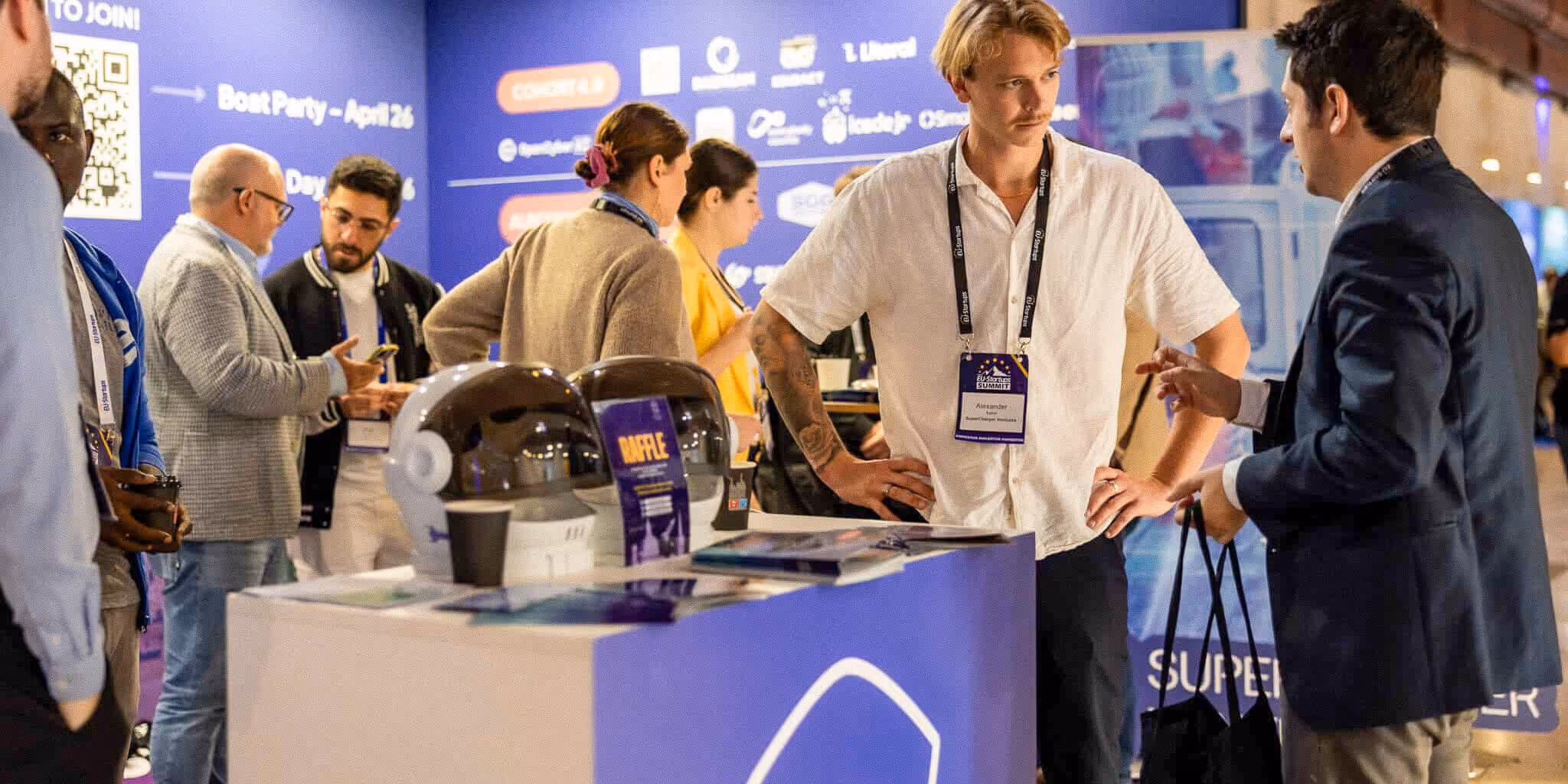People networking and discussing at a tech conference booth with promotional materials and helmets on display.