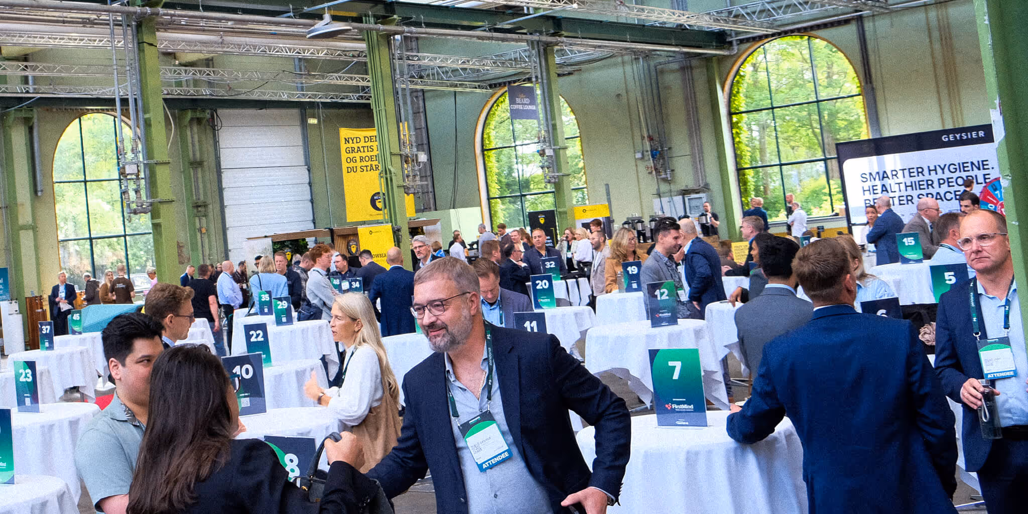Crowd of people networking and conversing at a professional indoor event with numbered tables and large arched windows.