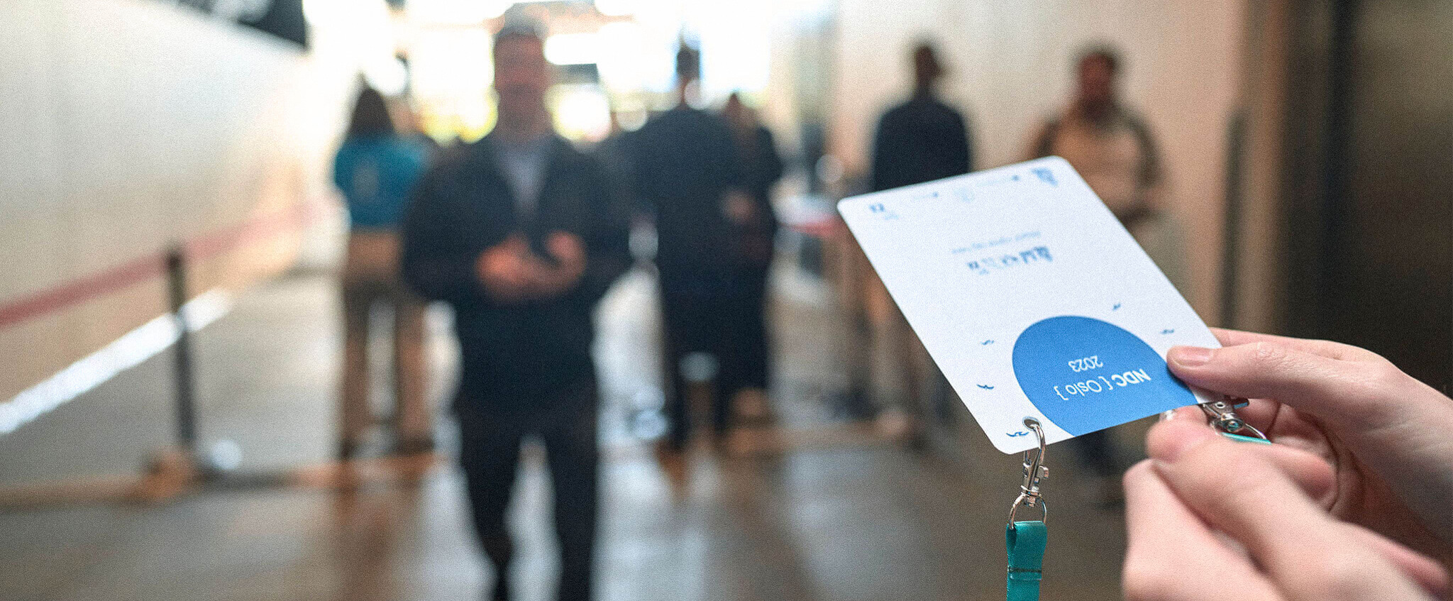 Close-up of hands holding an event badge with blurred people in the background at a check-in area.