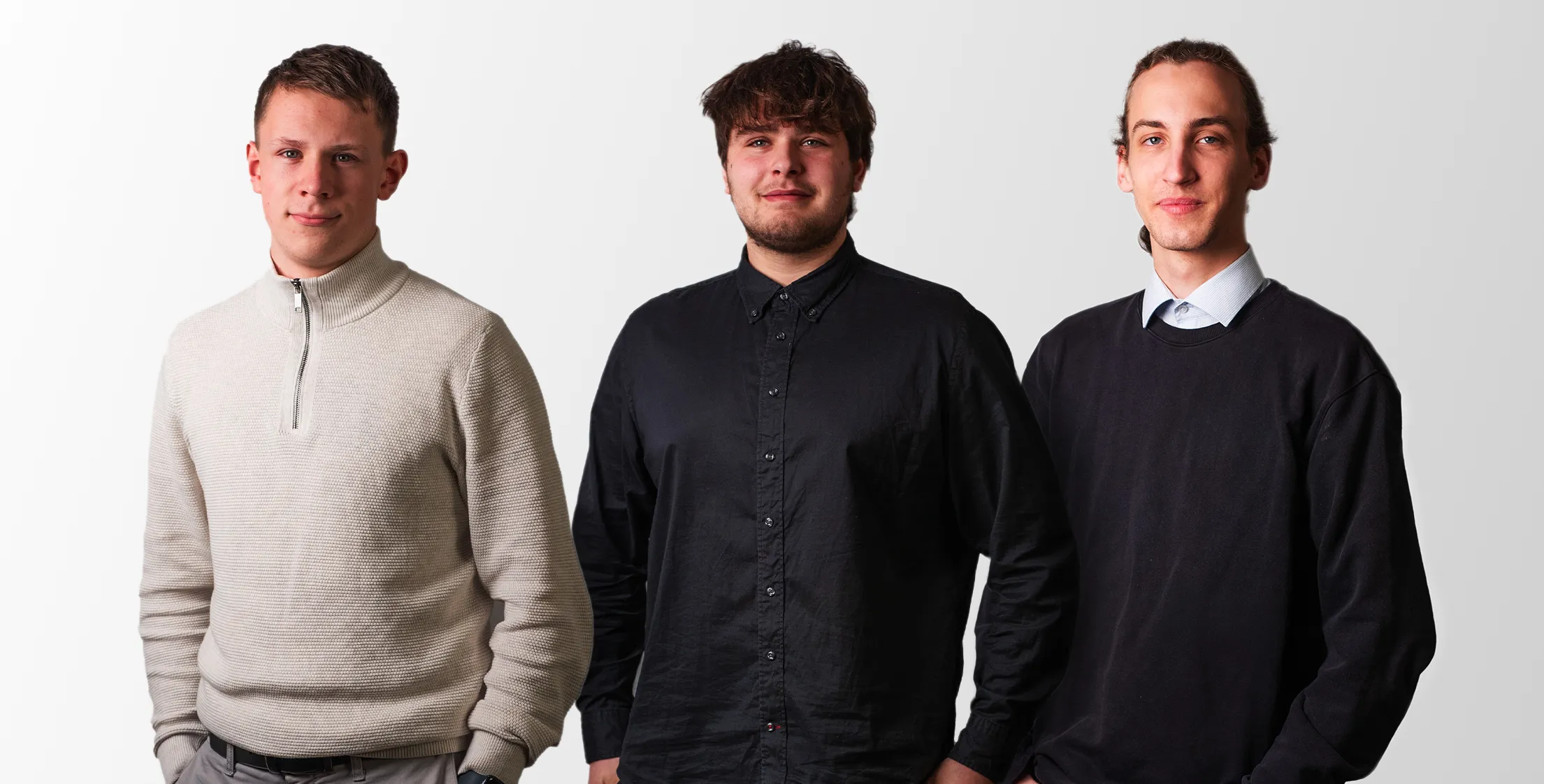 Drei junge Männer stehen vor einem weißen Hintergrund, der erste trägt einen hellen Pullover, der zweite ein schwarzes Hemd, der dritte einen schwarzen Pullover über einem hellblauen Hemd.