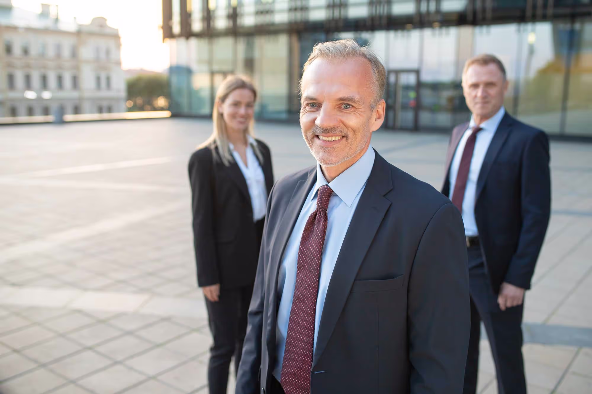 Three business professionals in suits standing outside a modern building, smiling at the camera.