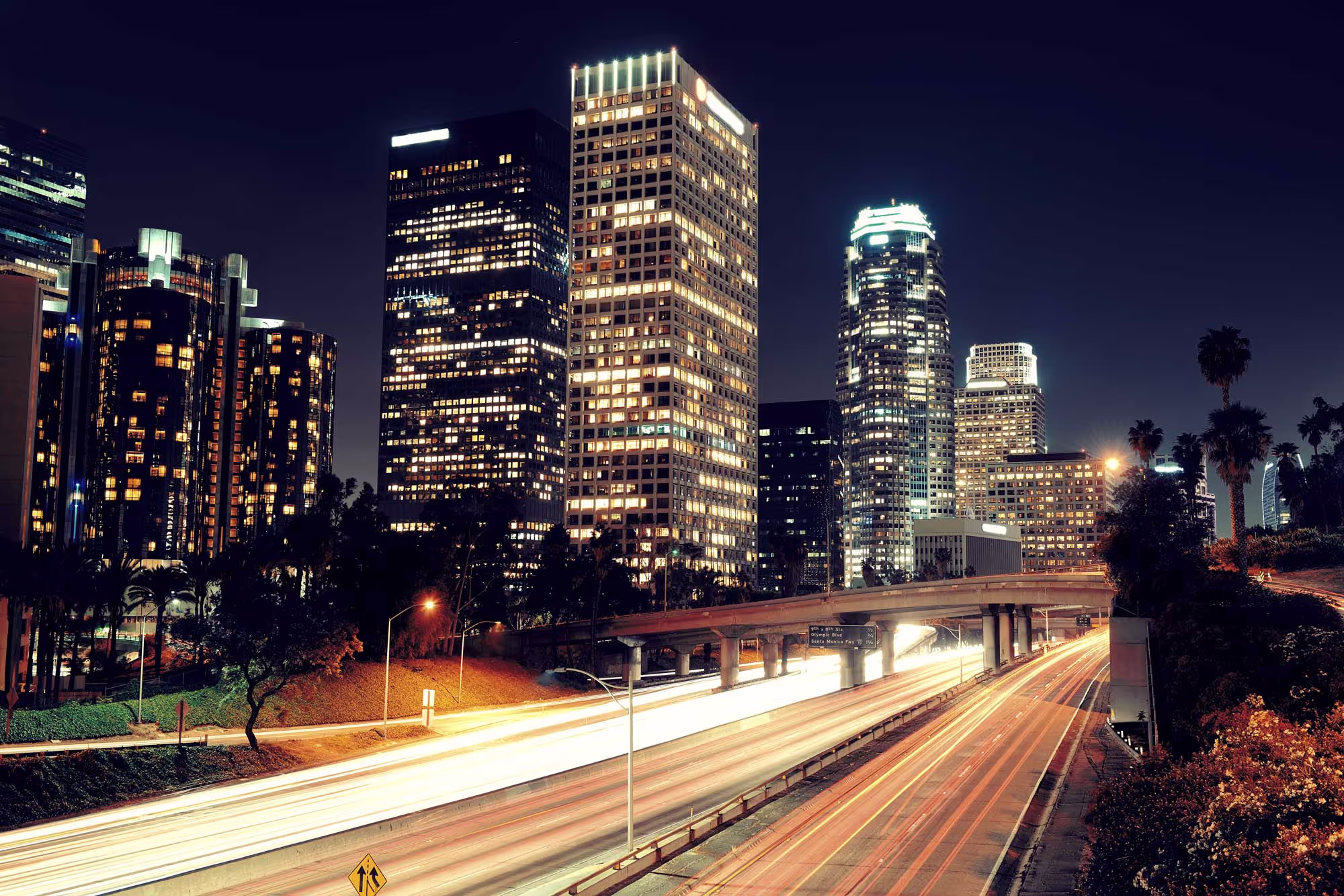 City skyline at night with brightly lit buildings and blurred highway traffic in the foreground.