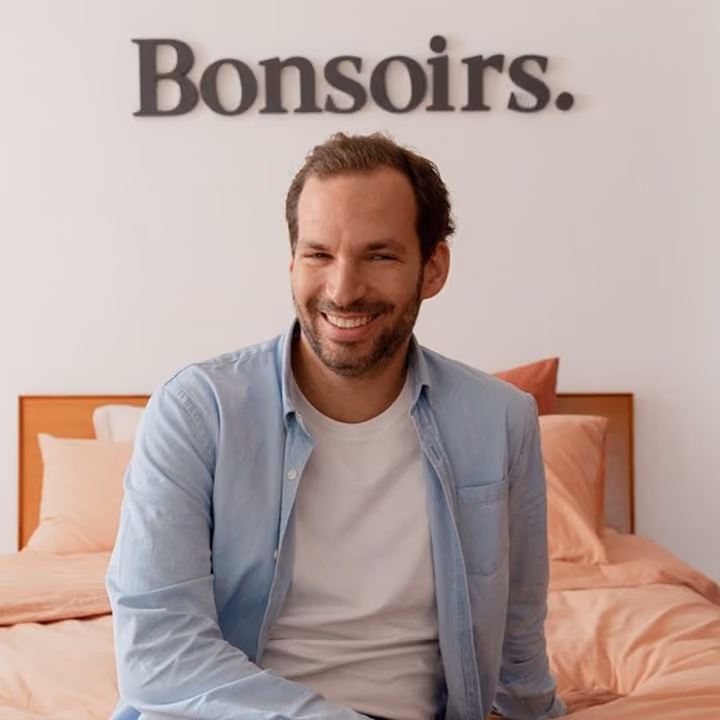 Un homme souriant portant une chemise bleue claire et un t-shirt blanc, assis sur un lit avec des draps roses, avec le mot « Bonsoirs. » inscrit en gros caractères noirs sur le mur blanc derrière lui.