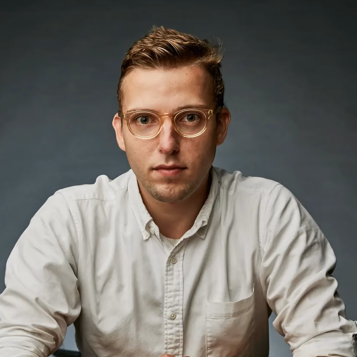 Jeune homme avec des lunettes transparentes et une chemise blanche regardant directement l'appareil photo sur un fond gris.