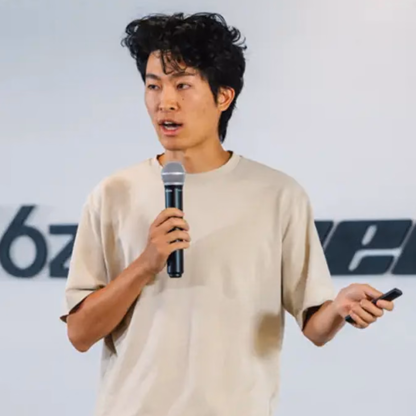 Jeune homme asiatique tenant un microphone et une télécommande, parlant devant un mur blanc avec un texte partiel en arrière-plan.