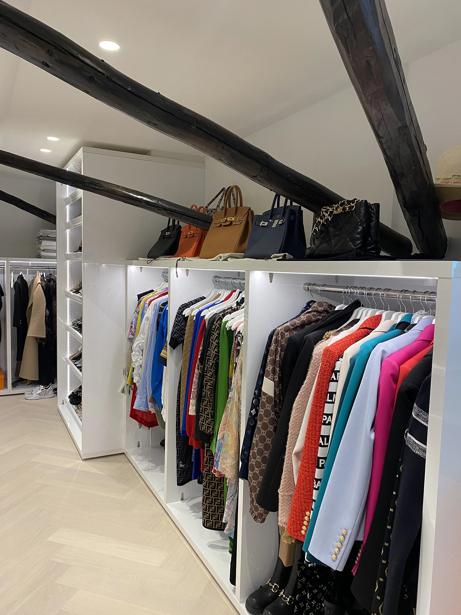 Stijlvolle inloopkast met op maat gemaakte witte kasten, open kledingrekken en ingebouwde LED-verlichting. Boven de rekken staan luxe designerhandtassen, terwijl de ruimte wordt gekenmerkt door donkere houten plafondbalken en een lichte visgraatvloer.