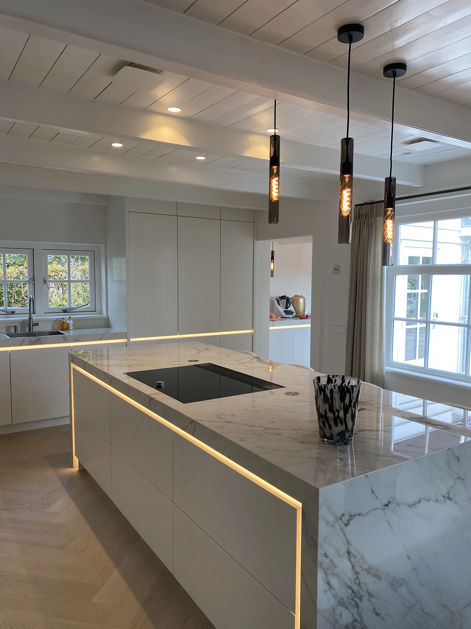 Moderne luxe keuken met een marmeren kookeiland, geïntegreerde LED-verlichting en een inductiekookplaat. De keuken heeft lichte houten kasten, een visgraatvloer en sfeervolle hanglampen met filamentverlichting.