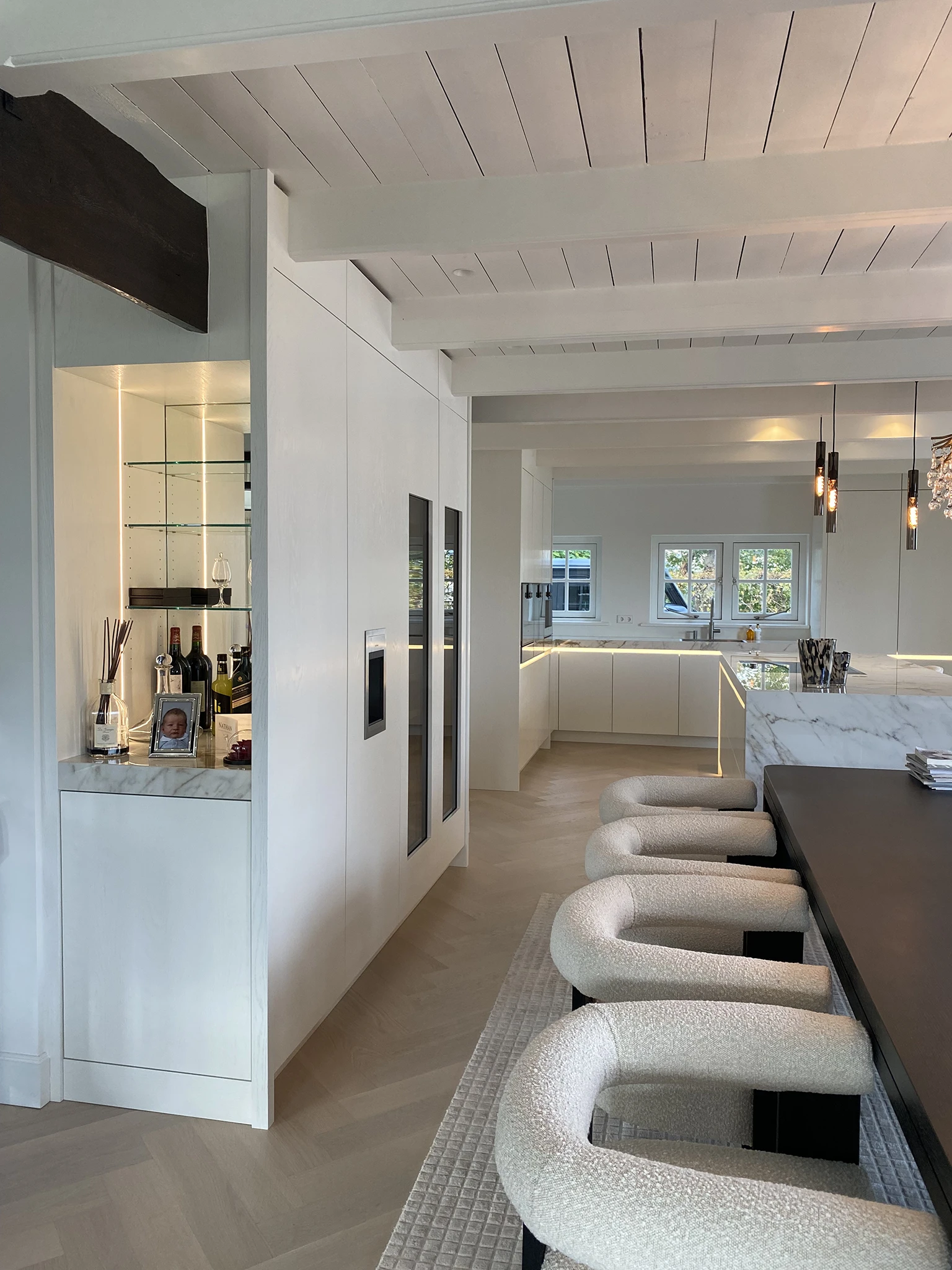 Stijlvolle open keuken en eetkamer met een marmeren bar, ingebouwde wijnklimaatkasten en een donkerhouten eettafel met bouclé stoelen. De ruimte heeft een lichte visgraatvloer, witte balkenplafonds en sfeervolle LED-verlichting.