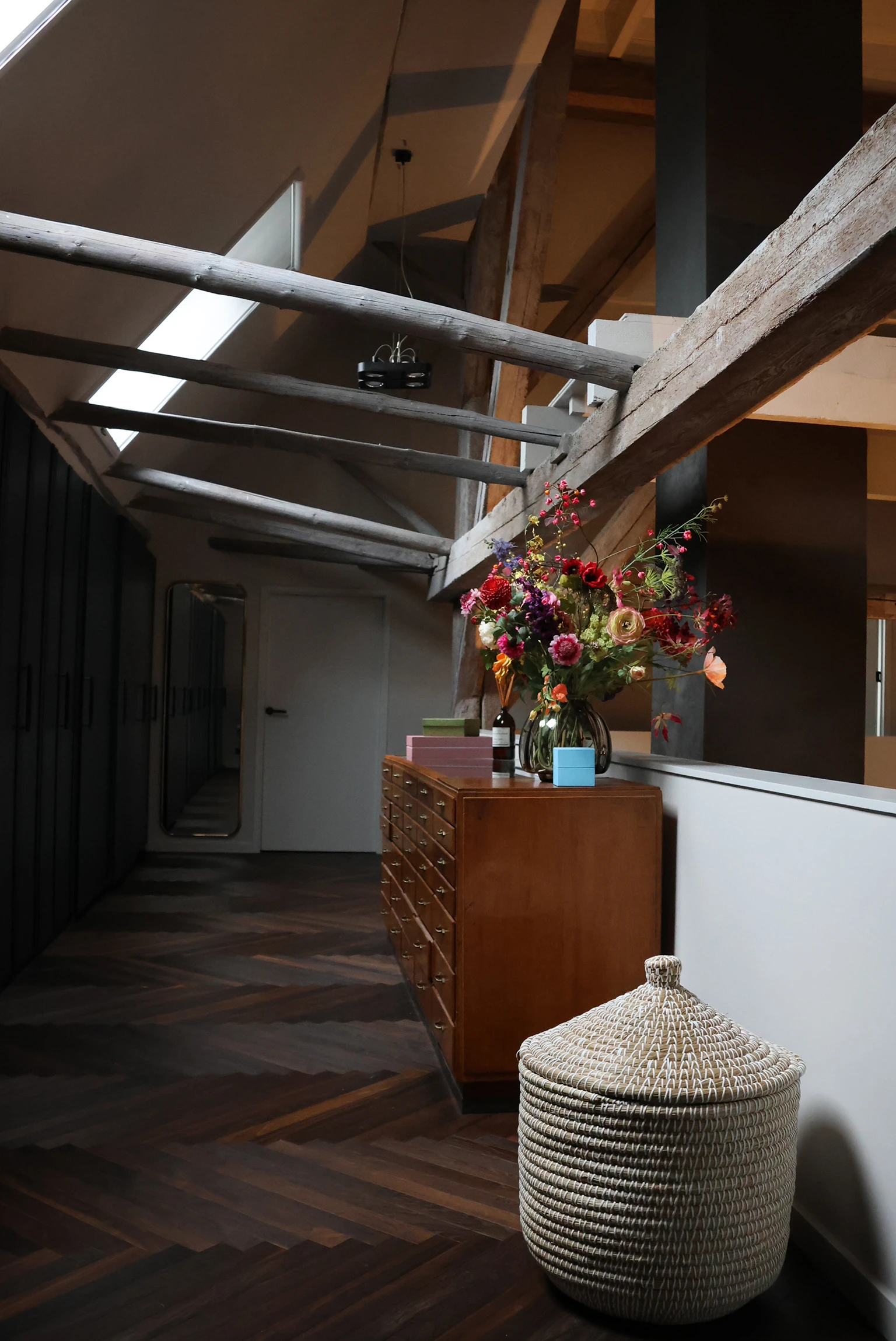 Sfeervolle bovenruimte in een gerenoveerde boerderij met houten balken, donkere visgraatvloer en een stijlvolle vintage kast.