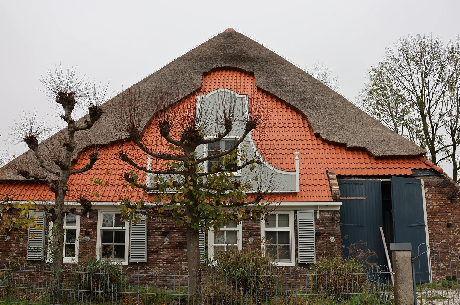 Gerestaureerde gevel van een boerderij met klassieke details, waaronder een karakteristieke klokgevel, houten luiken en een combinatie van een rieten en pannendak. De renovatie behoudt het authentieke karakter terwijl moderne afwerkingen zijn toegepast.