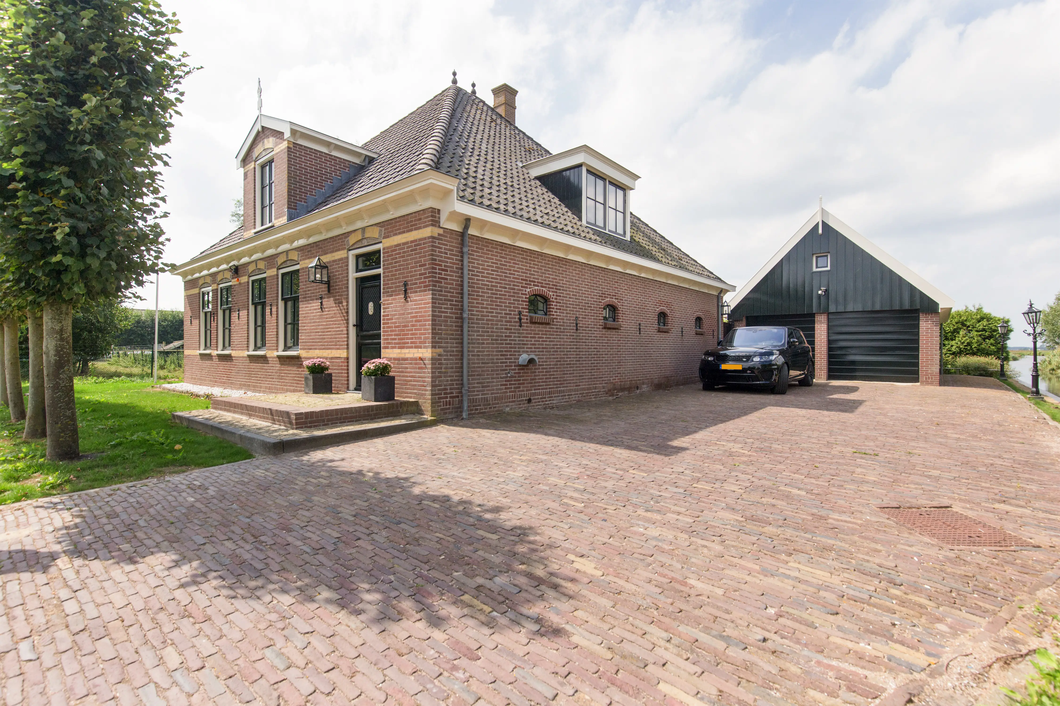 Gerestaureerde woonboerderij met klassieke rode bakstenen gevel, zwarte kozijnen en een bijpassende vrijstaande schuur. De woning heeft een authentieke uitstraling met een rieten kap en een sfeervolle, ruime oprit van oude klinkers.