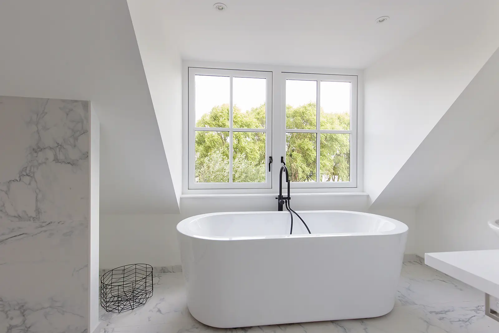 Een moderne, witte badkamer op een zolderverdieping met een vrijstaand bad onder een dakkapel met grote ramen die uitkijken op groene bomen.