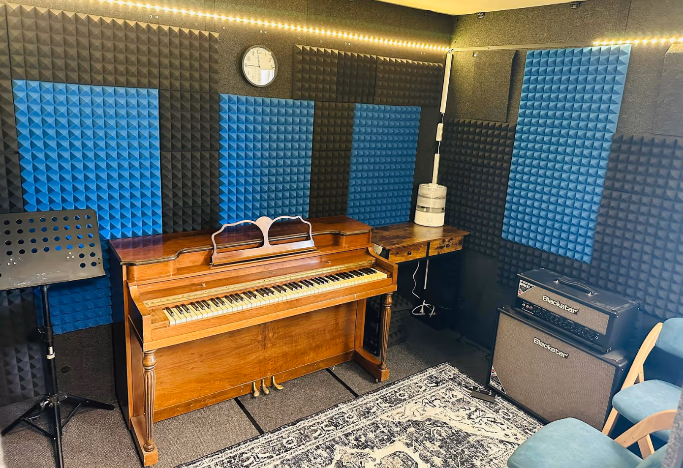 Interior of WhisperRoom 96120 S sound booth set up for music lessons with upright piano, amplifiers, music stand, acoustic panels, and LED lighting.