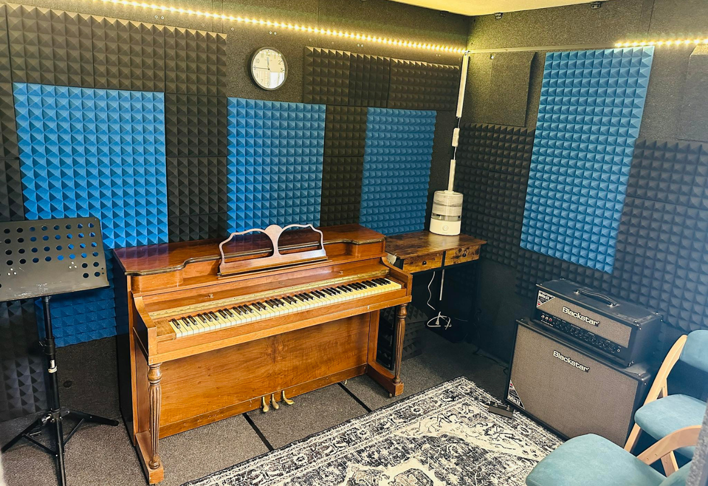 Interior of WhisperRoom 96120 S sound booth set up for music lessons with upright piano, amplifiers, music stand, acoustic panels, and LED lighting.
