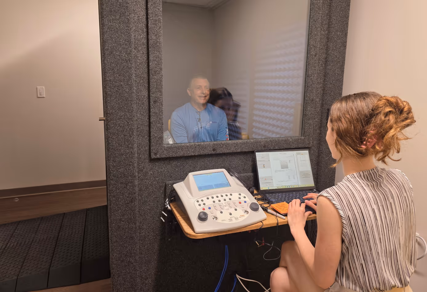 Audiologist conducting a hearing test in a WhisperRoom Audiology Basic Plus booth with wall window, desk, and testing equipment.