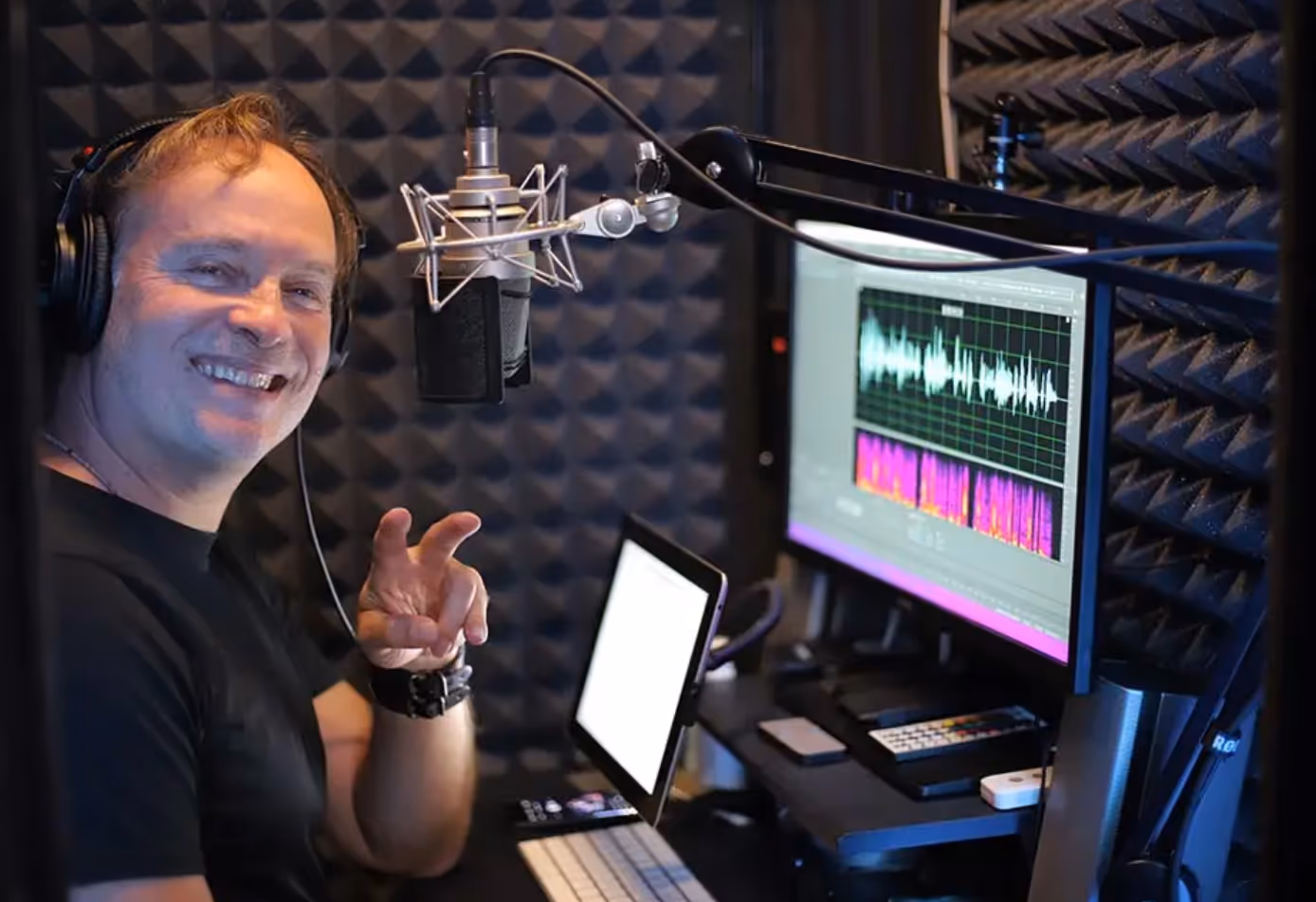 Voice actor recording inside a WhisperRoom MDL 4848 sound booth with microphone, headphones, acoustic foam, and computer workstation.